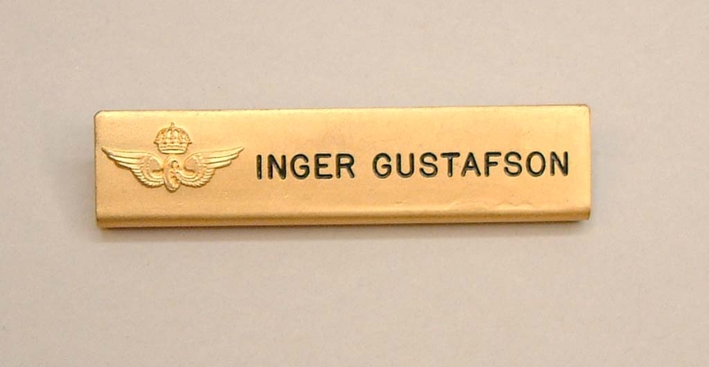 Namnbricka av gul metall, och bevingat hjul. Med SJ:s krönta vinghjul i svag relief, och namnet "Inger Gustafson" präglat i svart.