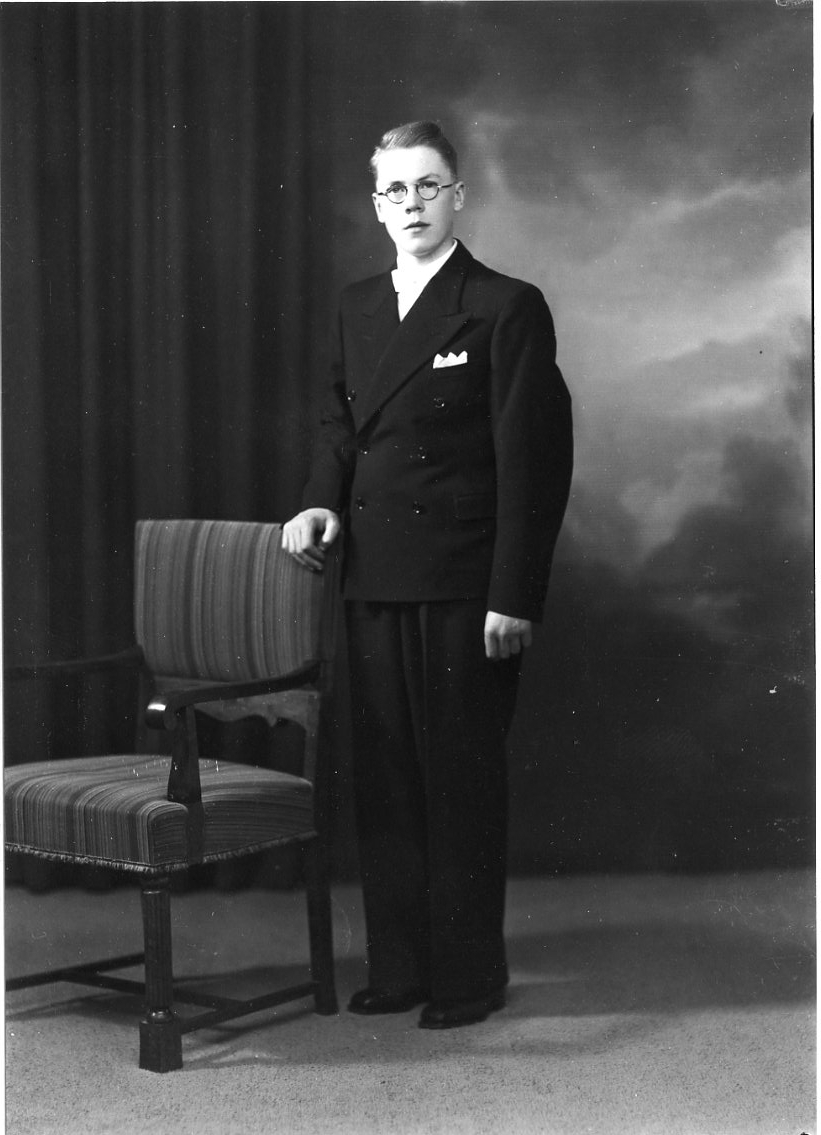 Porträtt av Bengt Ingesson från Örserum. En yngling i dubbelknäppt kostym, ljus skjorta och fluga. Näsduk i bröstfickan. Han står vid en stoppad stol.