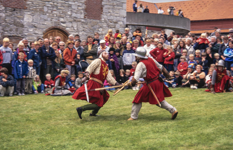 To riddere i rustning og med røde overkapper utkjemper blankvåpenkamp foran Storhamarlåven. Mellom ridderne og låven er det en stor menneskemengde som ser på kampen.