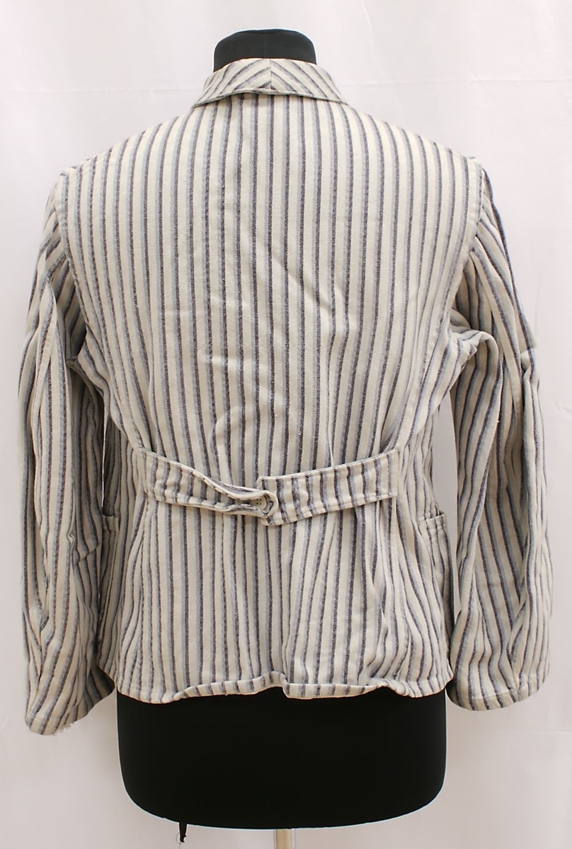 Enkel langermet bomullsskjorte med krave. Vertikalt stripemønster i blått og hvitt. To enkle lommer og fem knapper i front. Påsydd halvbelte med knepping bak. Knappene ser ut til å være laget av papp-liknende materiale, med hard overflate, muligens bakelitt.