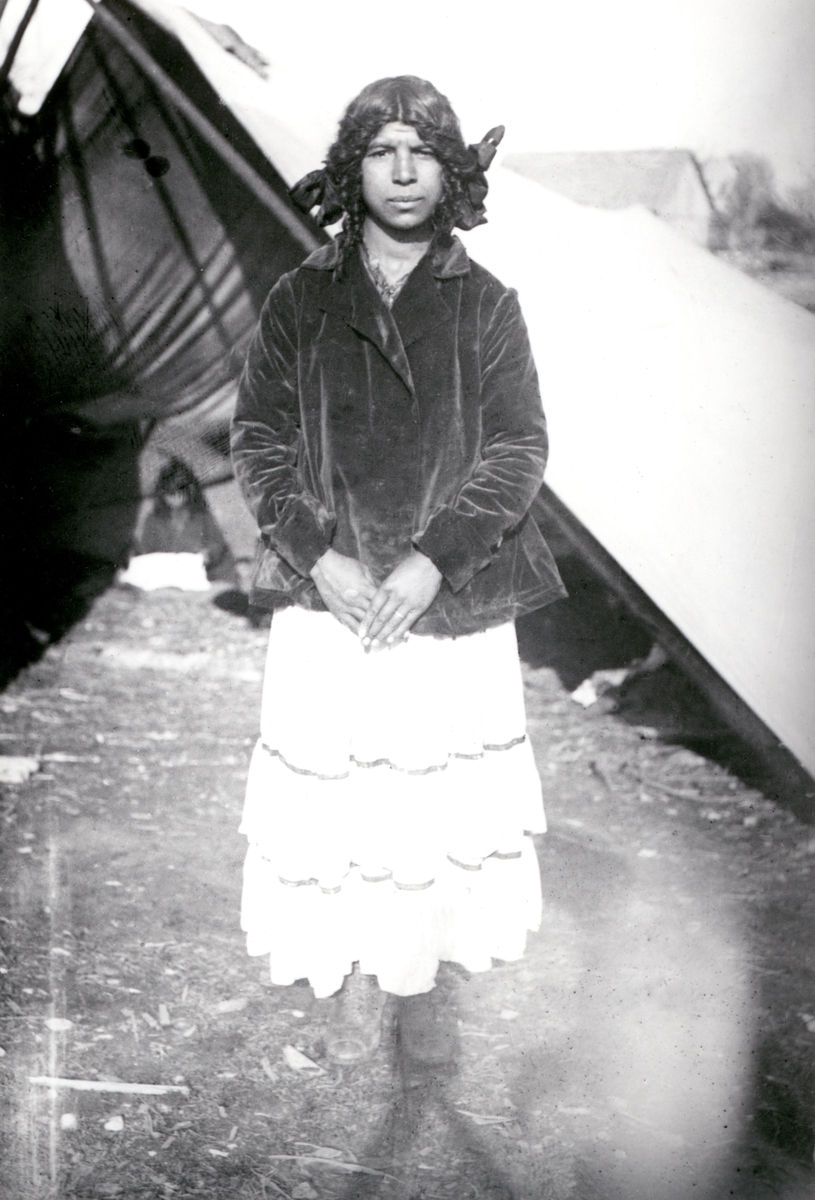En ung romsk kvinna står utanför ett tält, troligen tagen i Byxelkrok på Öland sommaren 1920. Kvinnan på bilden är antagligen ogift eftersom hon inte bär den romska sjaletten, dikhlon, som var brukligt för gifta romska kvinnor vid 1900-talets början.