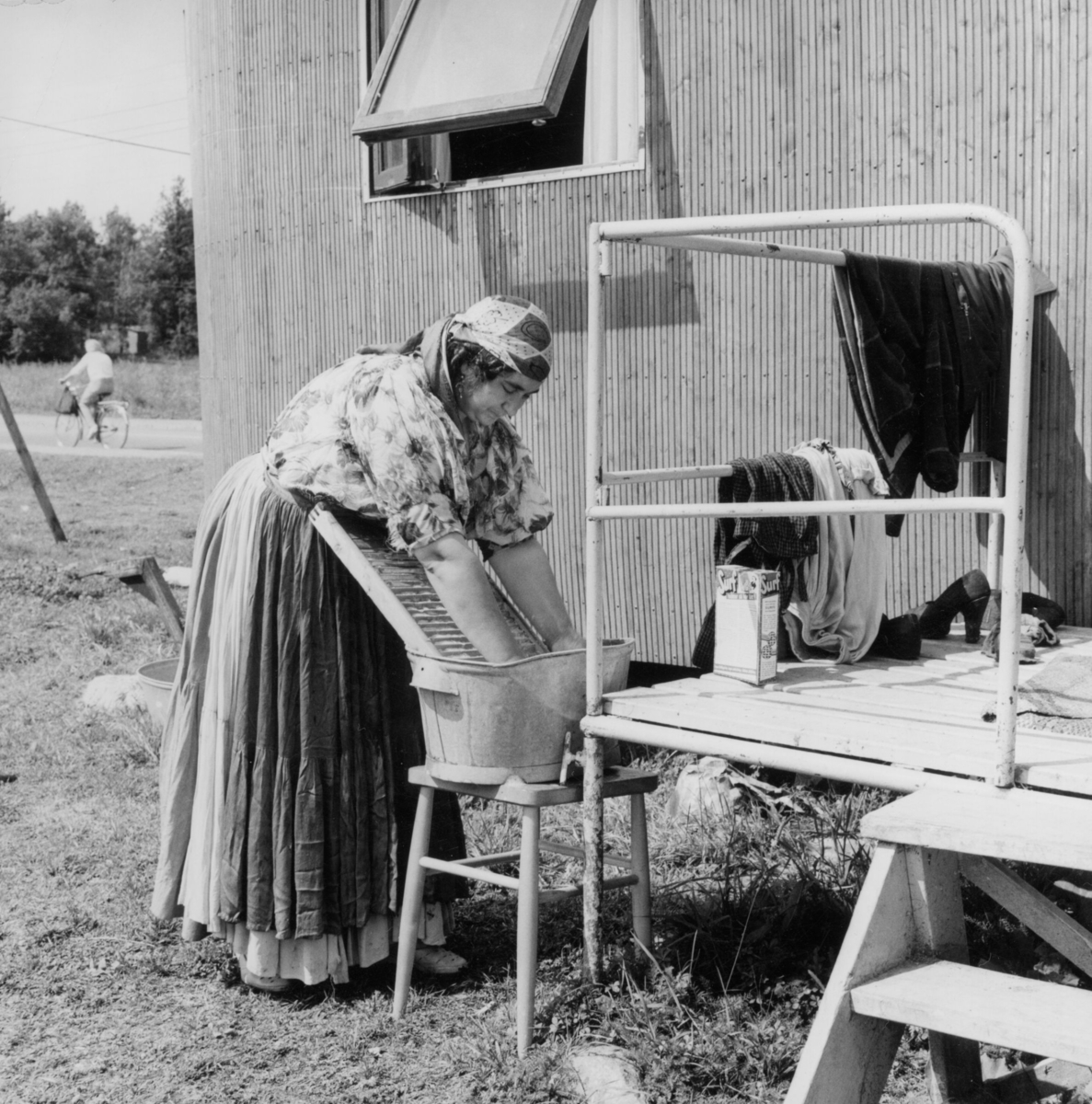 En romsk kvinnar tvättar utanför sin bostadsvagn i Tantolunden, Stockholm. Hon bär förkläde och en huvudduk.
