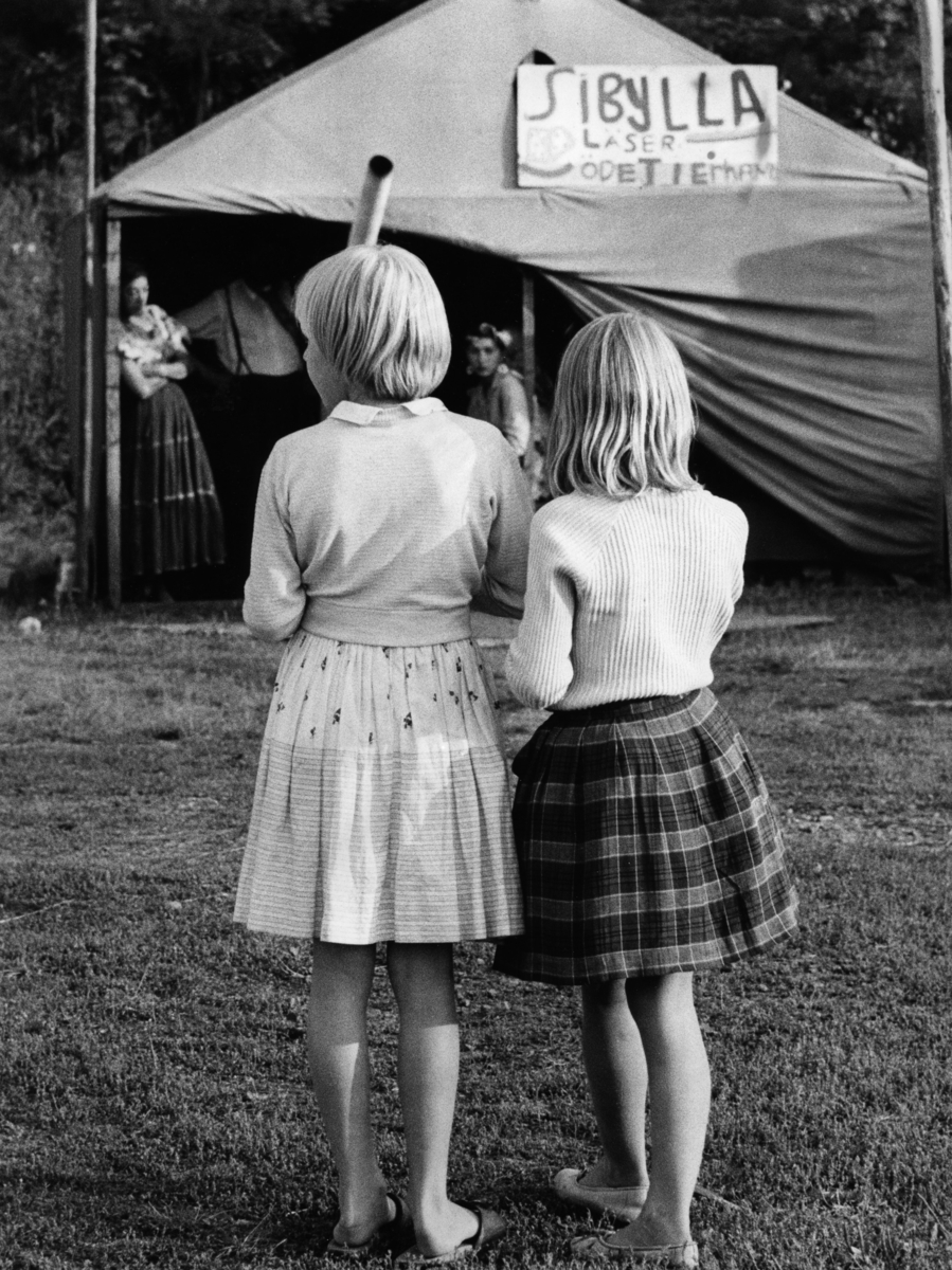 Två flickor tittar mot ett romskt tält. På tältet hänger en skylt med texten "Sibylla läser ödet i er hand".