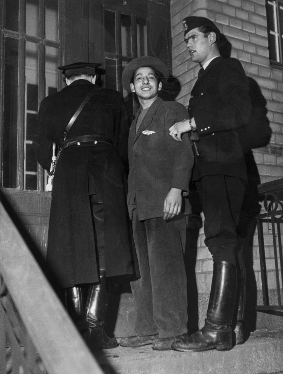 Två poliser eskorterar en man in på Enskede polisstation efter ett bråk i ett romskt läger vid Lilla Sköndal år 1950. Den anhållne mannen tittar glatt in i kameran.