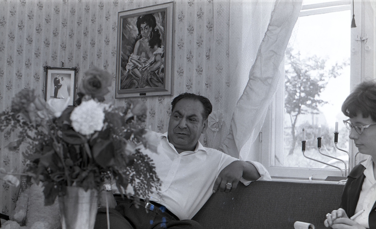 En man sitter i sitt vardagsrum och blir intervjuad av en journalist. Bilden är tagen i samband med ett nyhetsreportage för Skånska Dagbladet. Efter att de svenska romerna i Sverige erkändes som medborgare år 1952 uppstod debatt kring gruppens svåra levnadsförhållanden.