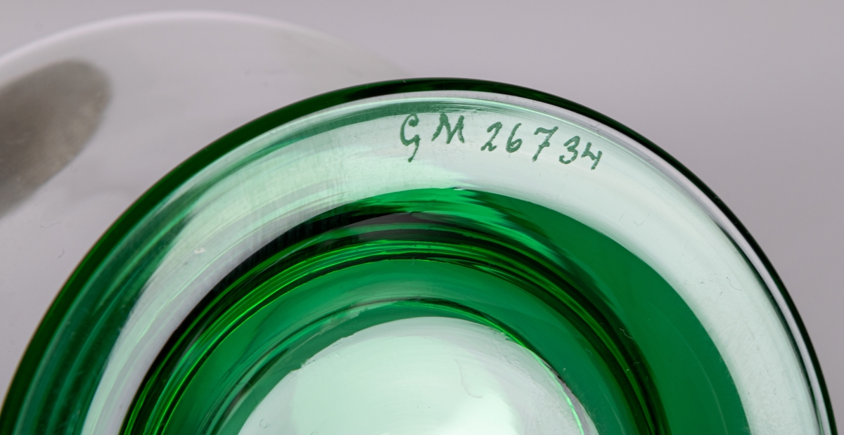 Vas av ofärgat glas på låg, utkragad fot i grönt glas. Delvis konisk med hög, smal hals. Design Gunnar Cyrén för Orrefors Glasbruk.