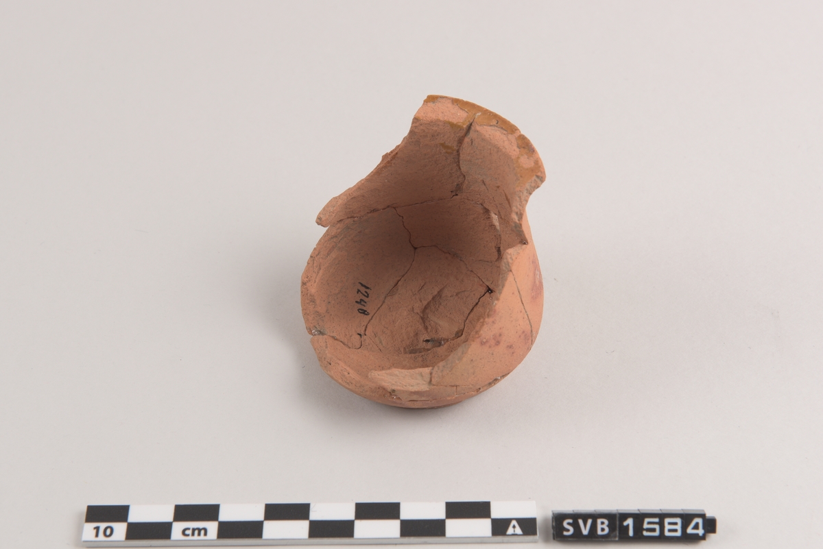 Terracotta-farget keramikk kopp. Bredere nedentil. En kant øverst. Koppen er ikke hel, men er limt sammen av fragmenter. Spor etter glasur.
