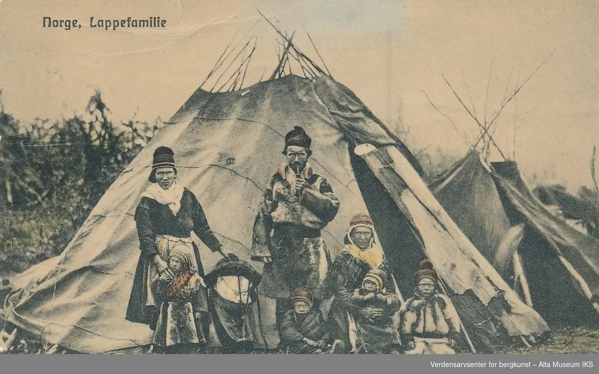 Samisk familie foran to lavvoer. Tre voksne og fire barn. Kvinnen til venstre holder en komse i venstre hånd.