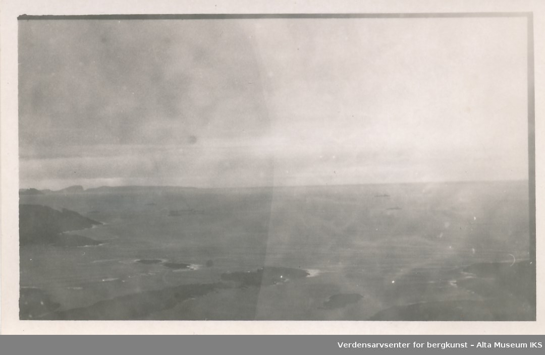 Motiv fra Finnmarkskysten. I forgrunnen og til venstre i bakgrunnen er det øyer og holmer.  På sjøen er det tre større og mindre fartøy.