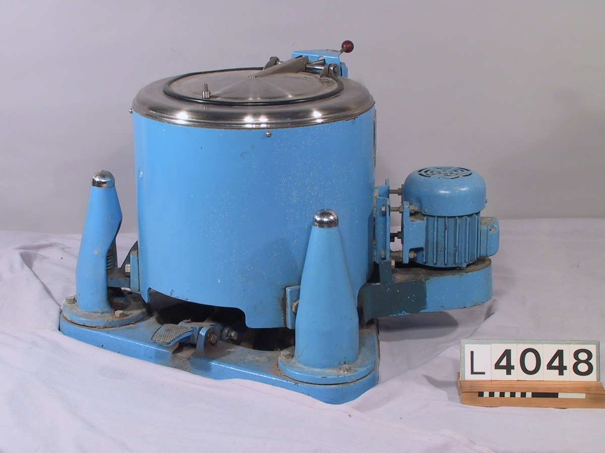 Fristående centrifug med tre fjäderben. Lock och topplåt i rostfritt stål. För övrigt är centrifugen blåmålad med bromspedal fram och motorn på sidan, bak.