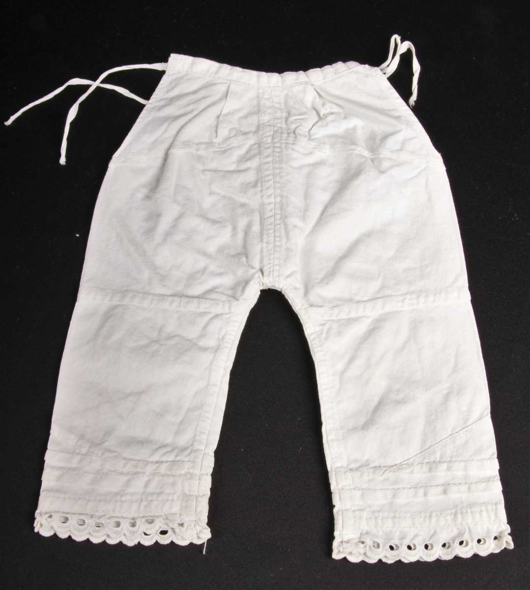 Hvit bomullsmamelukk til dukke eller spedbarn, med knyting i sidene og blondekant nederst på beina.