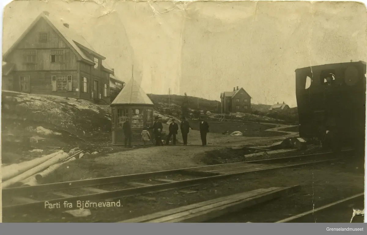 Parti fra Gruvebyen, Bjørnevatn ca 1915. Bak til venstre: Funksjonærbolig, i midten folk i kø utenfor Folkets hus` kiosk og i forkant et damplokomotiv på skinner.