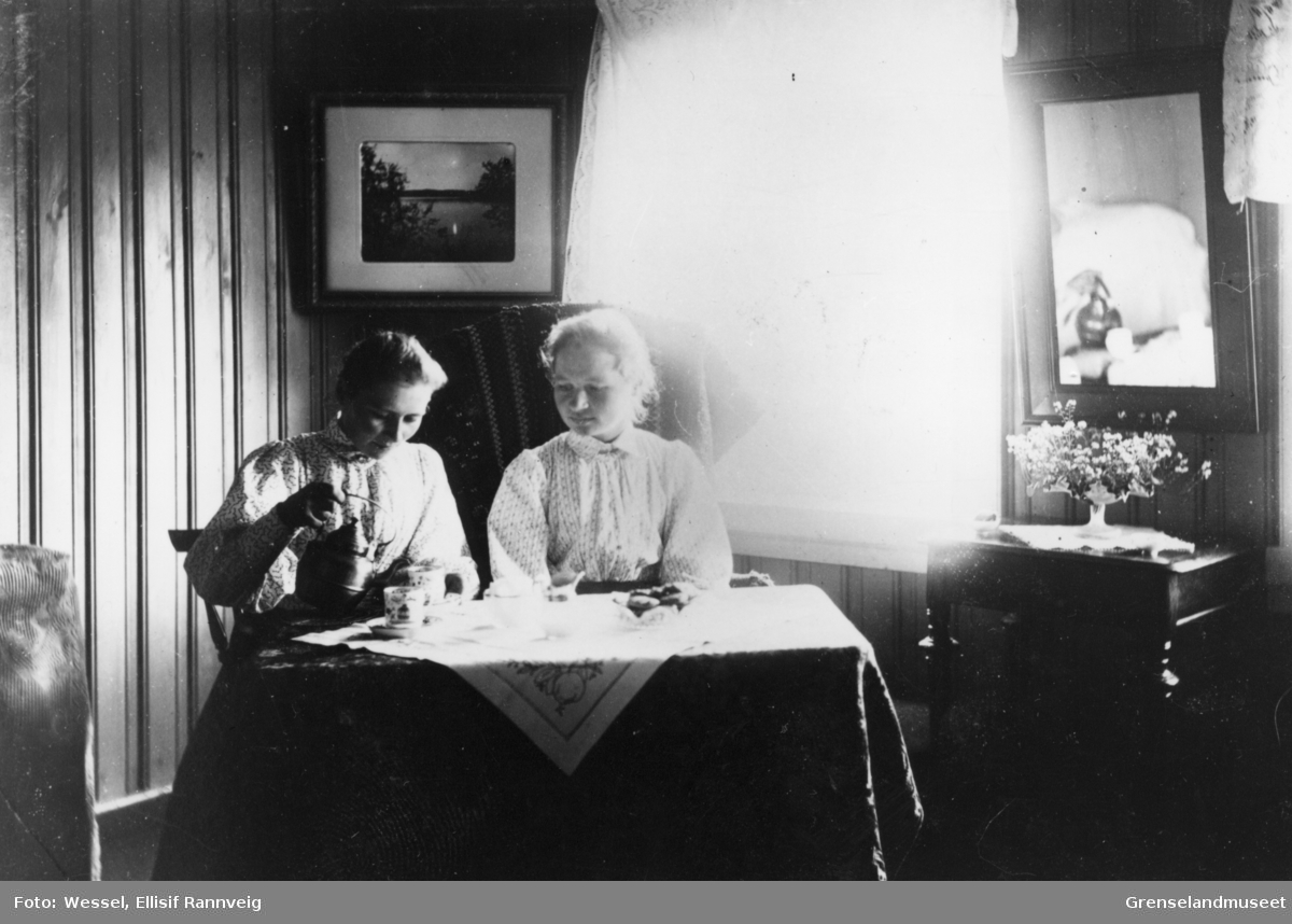 Søstrene Lilleng; lærerinne Elise og jordmor Annette, sitter ved et bord og drikker kaffe. Sannsynligvis er søstrene på besøk i Doktorgården Solheim, Kirkenes. På veggen henger et fotografi som Wessel selv har tatt.