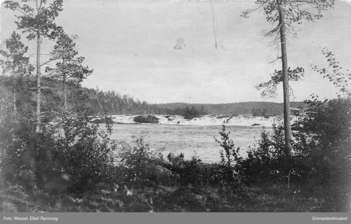 En elvebåt ligger på elvebredden i Pasvikelva, Skogfossen (Männikafossen) i bakgrunnen.