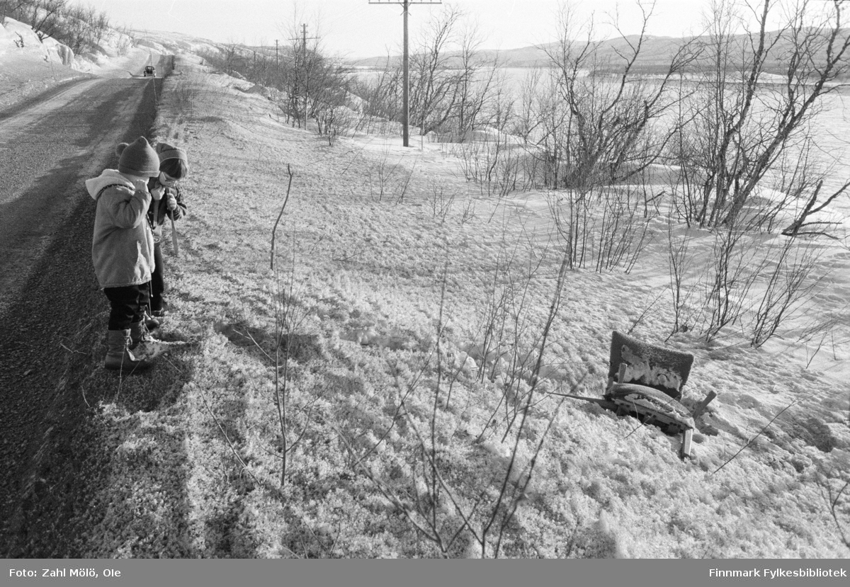 Vår i Tanadalen. To barn ved veikanten mellom Skiippagurra-Polmak. En lenestol er kastet på bakken i snøen. Tidspunkt er rundt 1970.