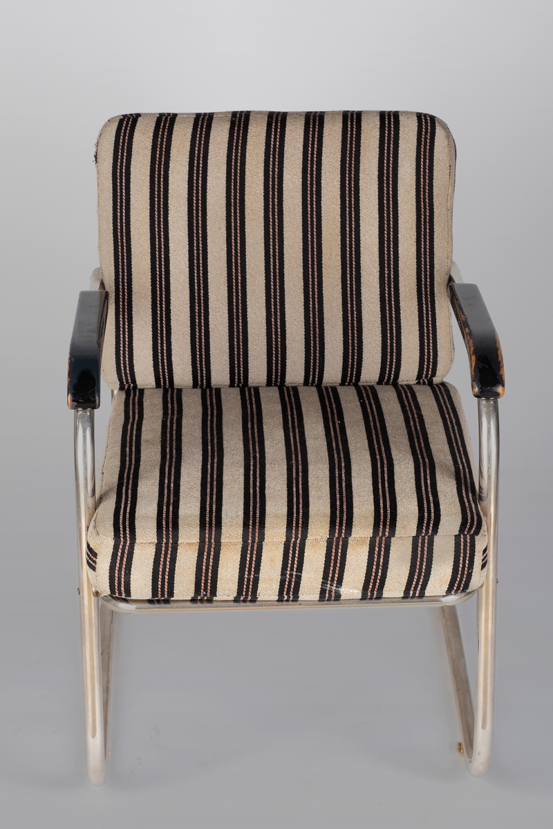 En stålrørslenestol med svartlakkerte armlener i tre. Stolen er satt sammen av to stålrørselementer. Ben og lener er ett element og sete og rygg er ett element. Festepunkt midt på rygg og foran på setet. Tre stålrørssprosser i ryggen og fjæring i sete. Gråbrunt møbeltrekk med vertikale brede, svarte striper som har to parallelle rader med smale vertikale striper. Disse er gråbrune med stiplete røde striper.