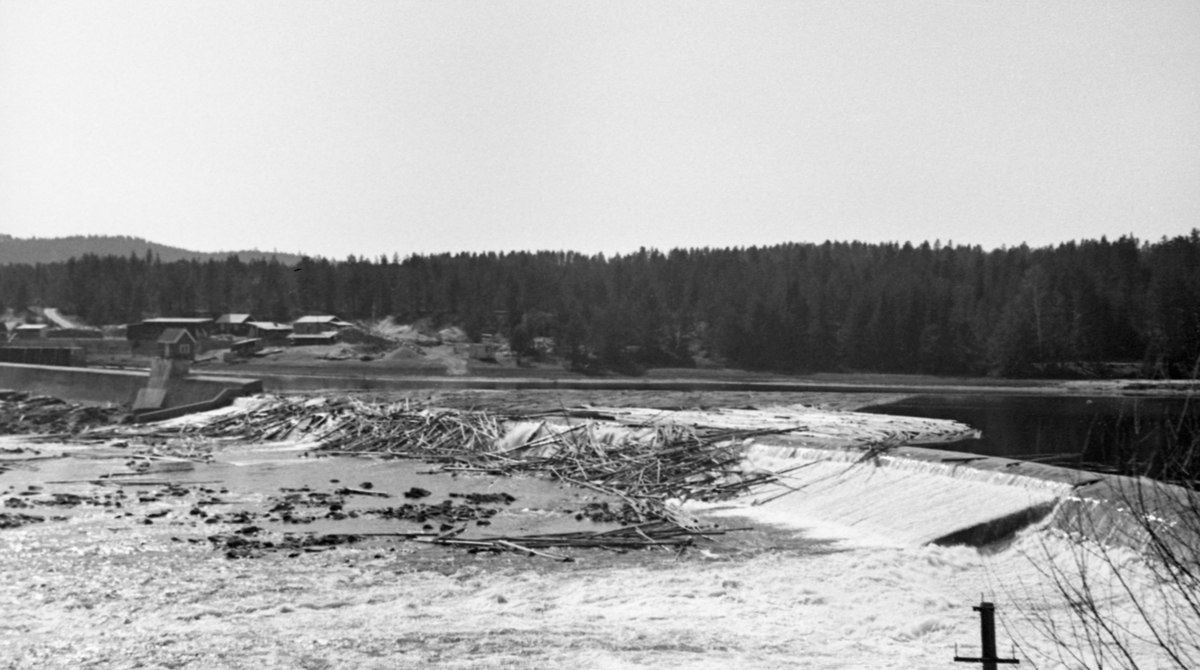 Skjefstadfossen i Glomma, nærmere bestemt i Heradsbygda i Elverum, Hedmark, fotografert i begynnelsen av mai 1944, på et tidspunkt da det var forholdsvis lite vann i elveløpet. Øverst i denne fossen hadde man i 1909-1910 bygd et elvekraftverk med en terskeldam som skulle stuve opp vann til turbinrøret. Overløpsdammen var 312 meter lang, utført i betong og støpt slik at damkrona var noe lavere i den østre enn i den vestre enden. Hensikten var å skape et «sug» i overflata som trakk fløtingstømmeret mot den østre delen av elveløpet. Av samme årsak brukte man å legge ei lense, et flytende stengsel land av sammenkjedete tømmerstokker, som skulle hindre at fløtingsvirket drev inn i inntaksbassenget for vann til kraftstasjonen, som lå i den vestre delen av elveløpet. Da dette fotografiet ble tatt var fløtinga for lengst i gang, men vannføringa hadde avtatt, slik at en god del tømmer hadde blitt liggende i fallsona på den vestre delen av overløpsdammen. Her var det for øvrig bygd en såkalt «tømmerløfter», et skråplan som skulle forebygge at stokkene ble splintret i nedfallssona under dammen.