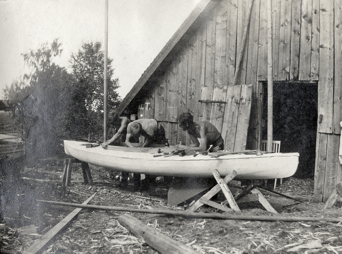 Några unga män sysslar med en segelbåt utanför en stor bod. 
Under fotot text: " - Båtvarvet - i Blädinge - ".