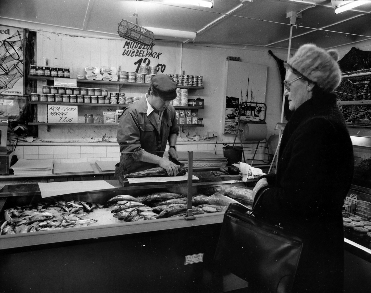 En man står bakom disken och paketerar en fisk medan en kvinna står framför disken och väntar. Västkustens fiskaffär, kv Pärlan. Fastigheten revs i månadsskiftet juli - augusti 1984.