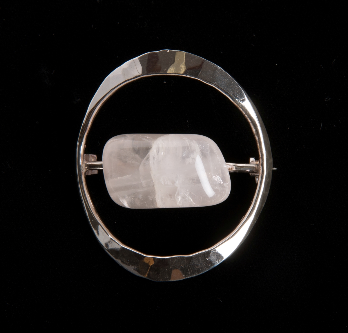 Åpen flathamret ring. En sølvstang er tredd i ringen som det er festet en stein på. Nålfeste bak stangen. Hvit, rektangulær stein. Ringen er hamret i to plan, 2+2 sider.