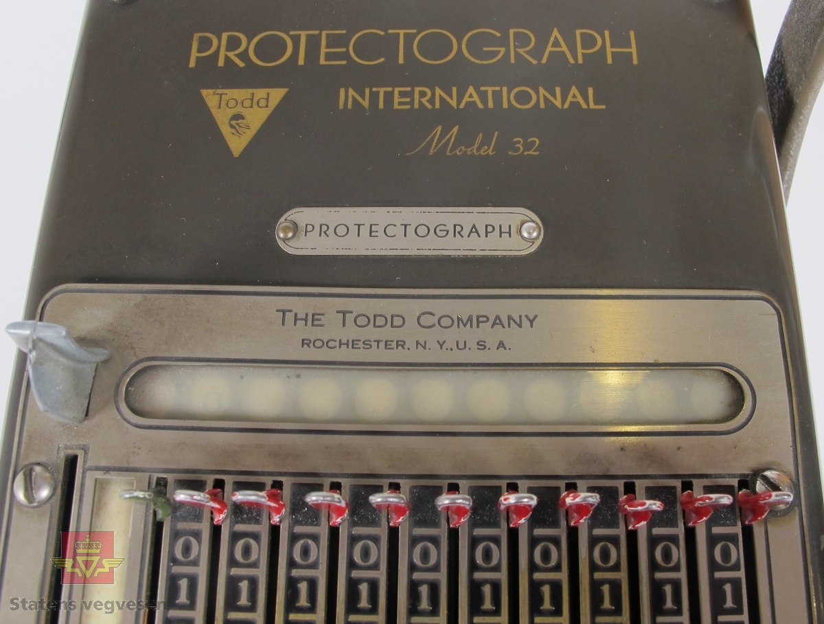 Protektograf i metall med påskriften PROTECTOGRAPH INTERNATIONAL Model 32 THE TODD COMPANY ROCHESTER.N.Y.,U.S.A. Har serienummer D320Z1229 montert på en metallplate bak på maskinen. Den har to sveivehåndtak, og betjeningspanel for innstilling av siffer. Den er i fungerende tilstand og stempler H.E-H.A/S før KR og sum. Teksten er svart og sifrene er røde. Mulighet for å stille inn flere typer valuta.