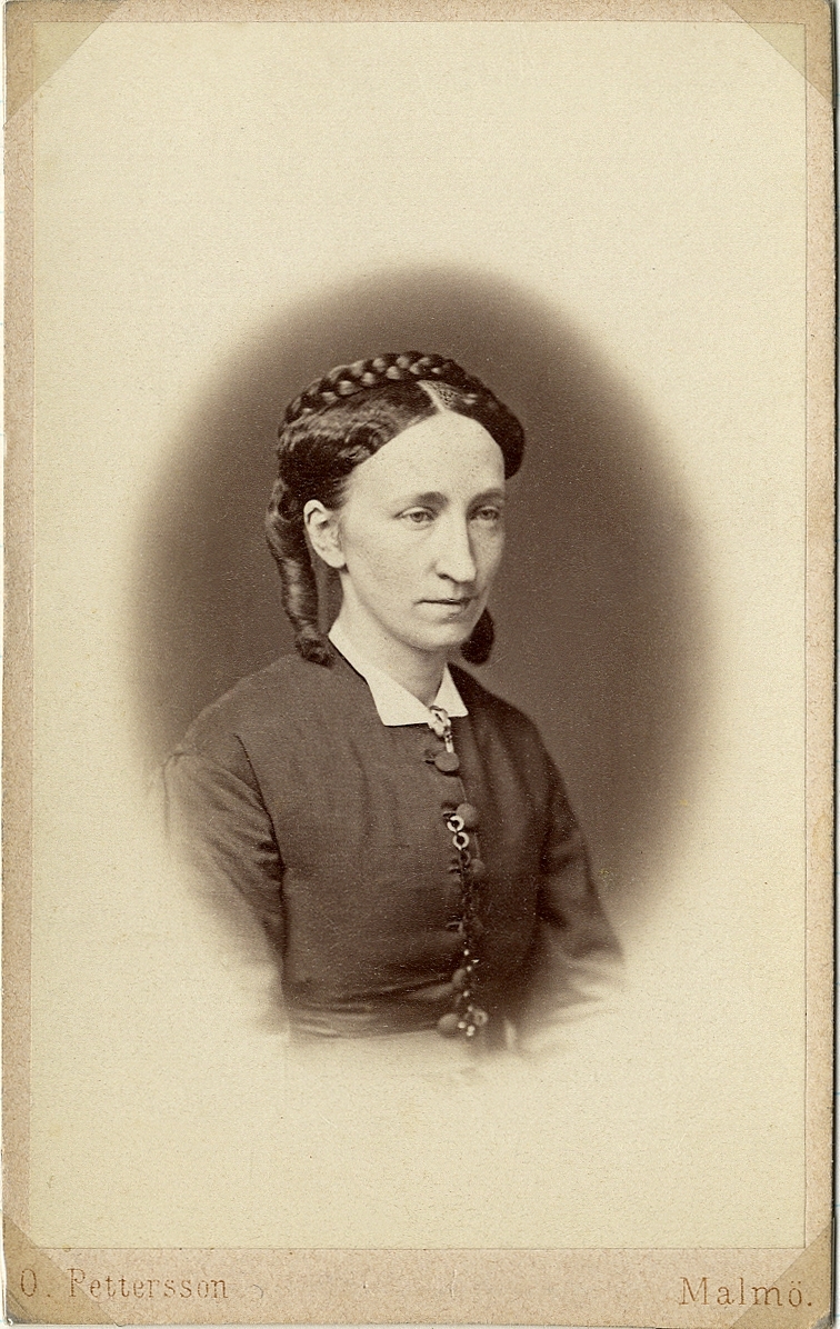 Porträttfoto av en kvinna i mörk klänning med enkel vit krage och ett halsband runt halsen. 
Midjebild, halvprofil. Ateljéfoto.