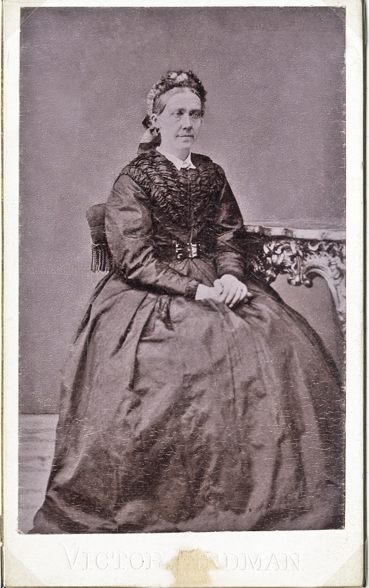 Porträttfoto av en äldre kvinna i mörk klänning med liten vit krage. I håret en hårklädsel med band och spetsar. Hon sitter på en stoppad stol med fransar, vid ett bord i nyrokoko. 
Helfigur, halvprofil. Ateljéfoto.

Troligen reprofoto från ett äldre foto (ca 1870) med okänd fotograf.