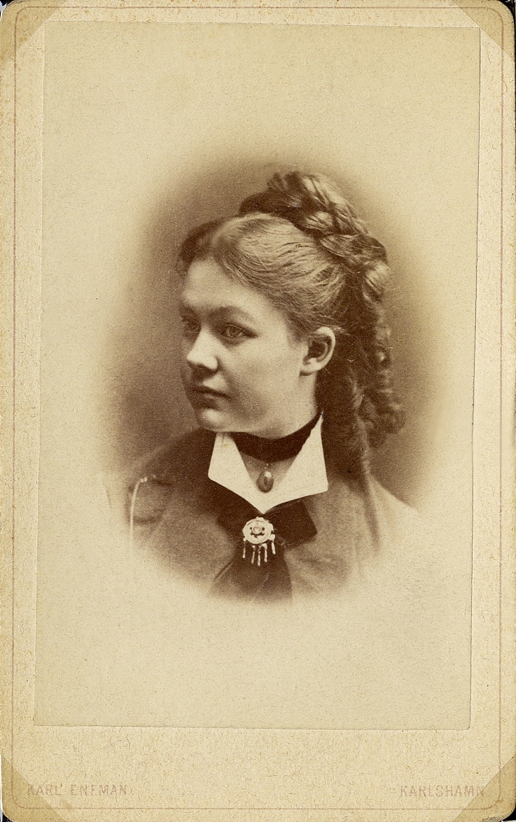 Porträttfoto av en kvinna i mörk klänning med vit krage. Vid kragen syns en brosch på en mörk scarf. Runt halsen har hon ett sammetsband med en berlock. 
Bröstbild, halvprofil. Ateljéfoto.