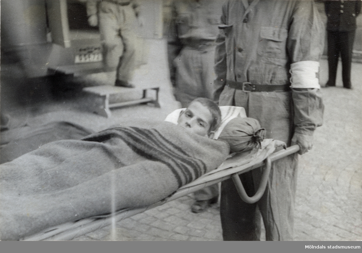 "Karantänsförläggning" på Kvarnbyskolan för överlevande från koncentrationsläger i Tyskland och Polen 1945.  
Skolan fungerade under denna sommar som beredsskapssjukhus.

En ung man blir buren på bår av Röda Korsets personal.