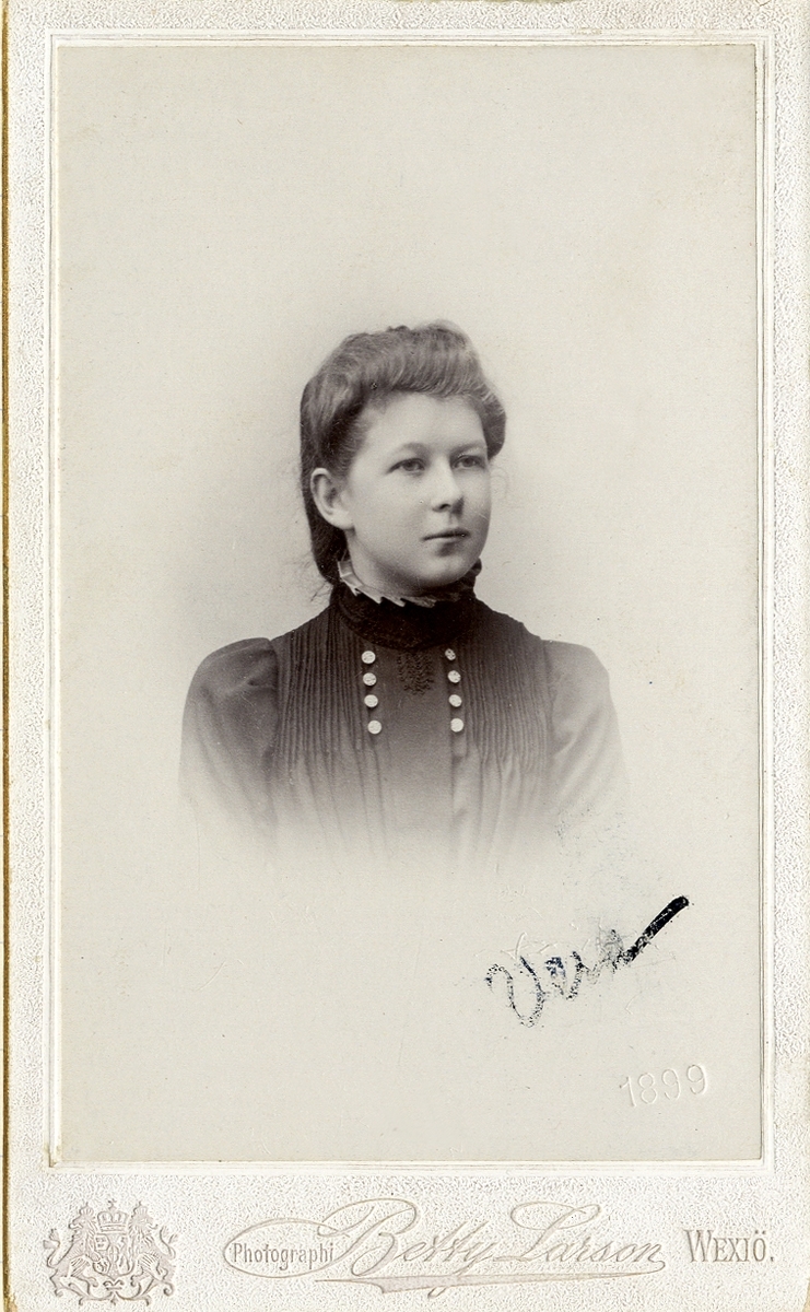 Foto av en ung kvinna i mörk klänning med hög krage och stråveck, Vid kragen syns två rader med prydnadsknappar. 
I nedre högra hörnet syns en autograf: "Vera" (på snedden). Där syns också ett inpräglat årtal: "1899". 
Bröstbild, halvprofil. Ateljéfoto.