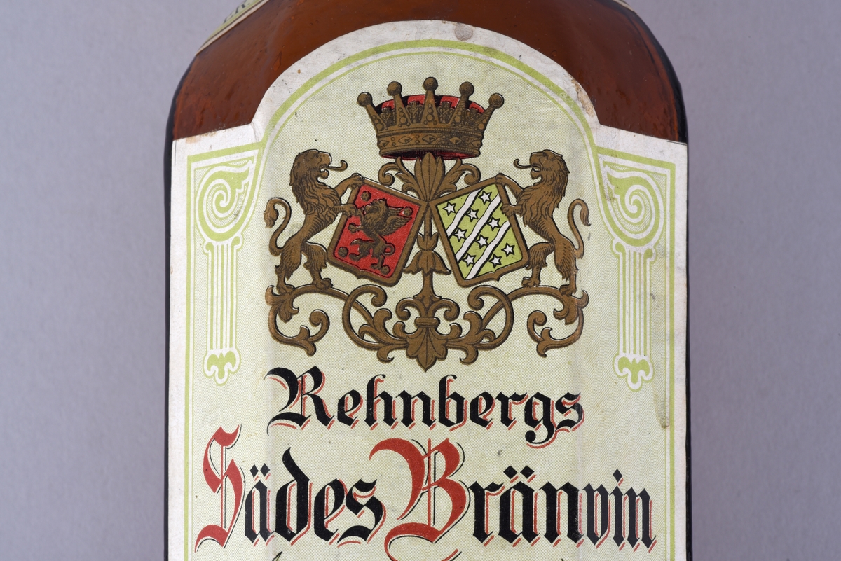 Åttkantig brun glasflaska för brännvin med etikett. Etikett för Rehnbergs sädesbrännvin, Söderköping. Ovanför texten på etiketten två motställda lejon som håller varsin vapensköld, den vänstra Östergötlands, den högra Söderköpings. Ovanför dessa en öppen krona.