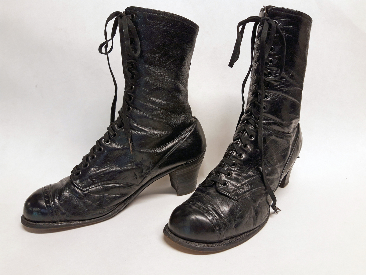 Et par sko i eske. Skoene har høye hæler bestående av skiver av lær lagt oppå hverandre og naglet i bunnen. Såla er av lær og er sydd på. Skoene er av svartfarget lær med kypertvevd bomullsstoff til fôr. Det er knyting på skoene. 