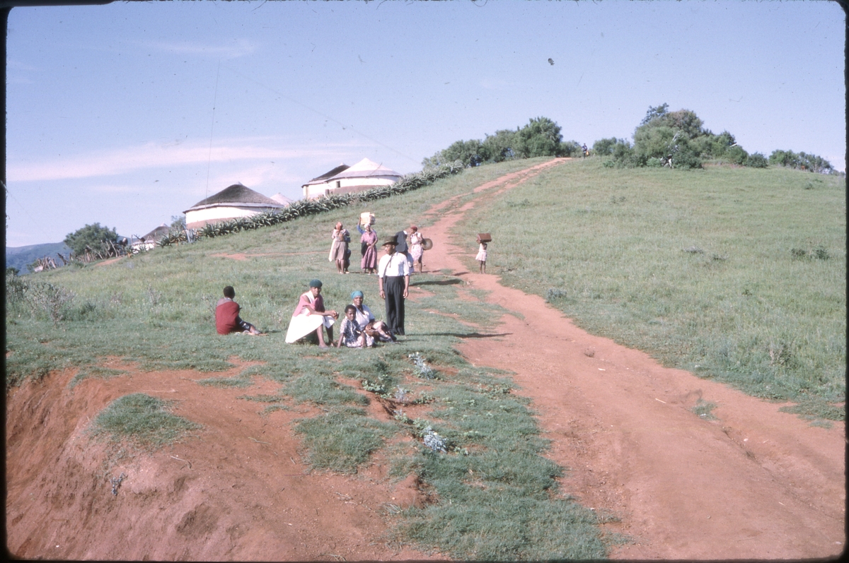 Mennesker stående og sittende langs jordvei på en åsside, med bebyggelse i bakgrunnen. 'Sagafjord' Around The World via Africa Cruise 1966.