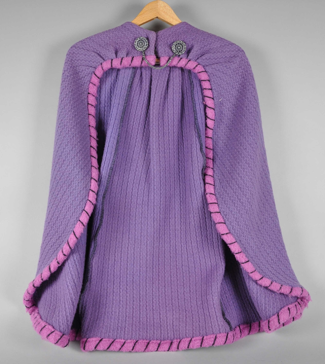 Lilla cape vevet av ull, med rosa kanter med svarte store sting. Foran på kragen lukkes kappen med to metallspenner med lenke mellom, og trykknapper.