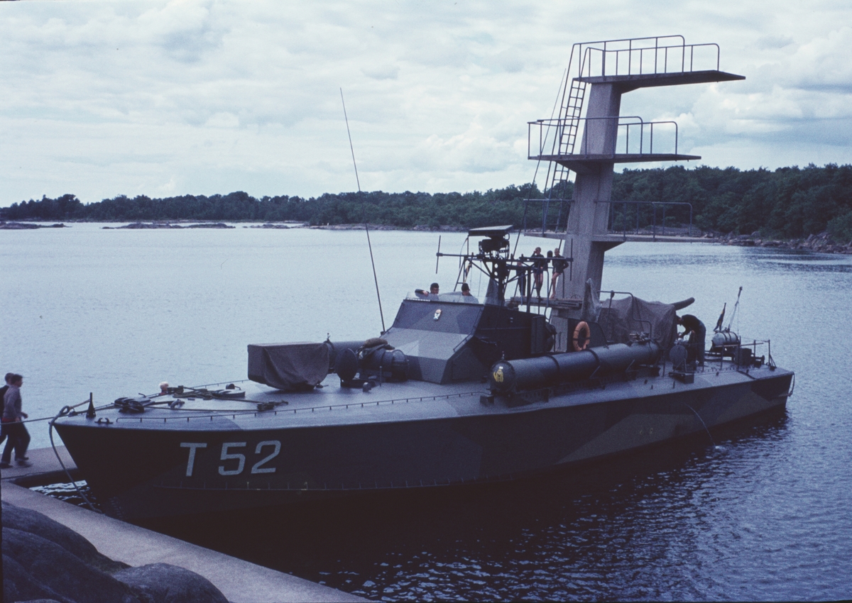 Bilden visar motortorpedbåten T 52 som ligger förtöjt på en brygga. Bakom den finns ett hopptorn och man ser en skärgårdslandskap.