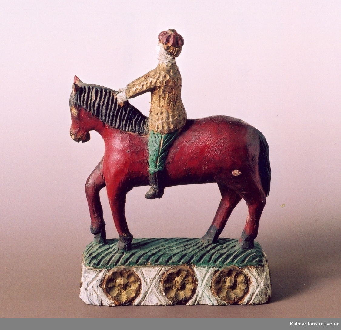 KLM 12927. Skulptur av trä. Ryttare och häst. Målad i starka färger, ryttaren i guldfärgad jacka och gröna byxor, hästen röd med brun man och svans, skulpturfoten dekorerad med en rad guldmålade öländska solvändor. Tillverkaren JAGS kallade skulpturtypen för Faraos ryttare. Tillverkad av träsnidaren Johan August Gustafsson, f 1852, d 1932, i Seby.