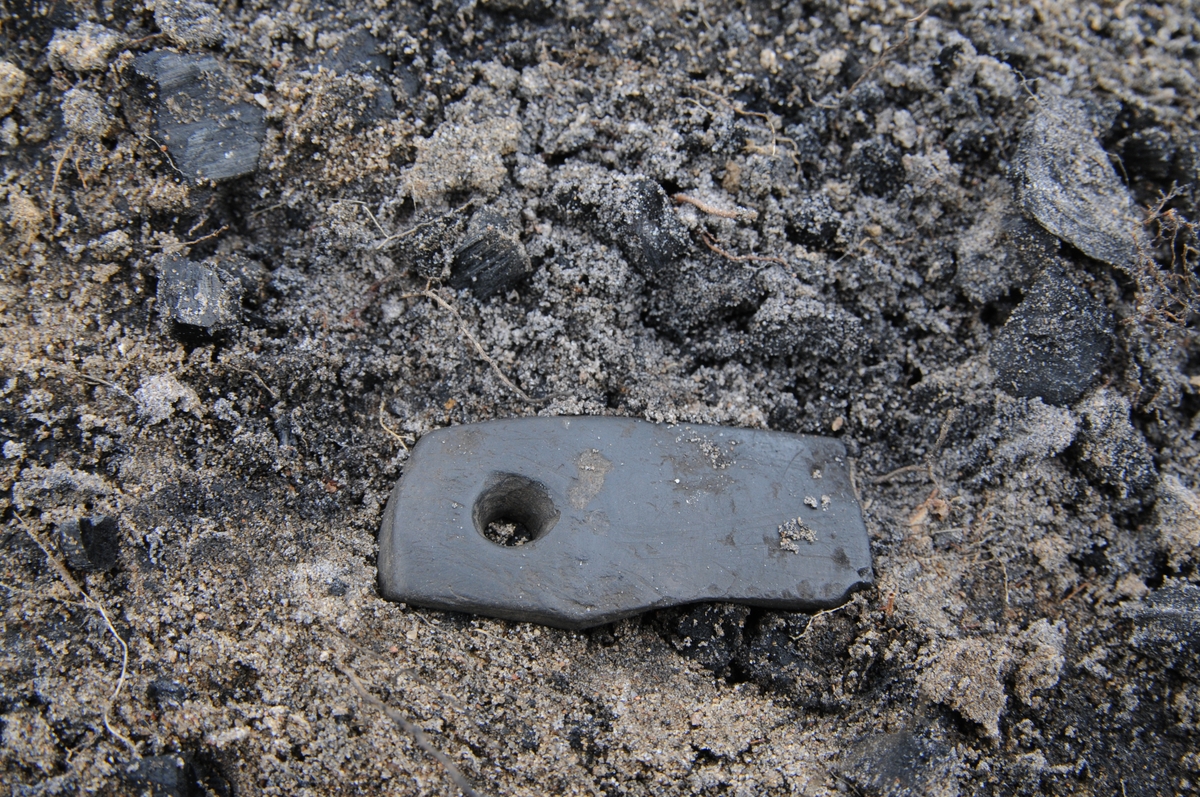 Ett bryne i skiffer med borrat hål. Påträffad i en gravhög vid en arkeologisk undersökning i Smålandsstenar i Villstad socken i Gislaveds kommun.