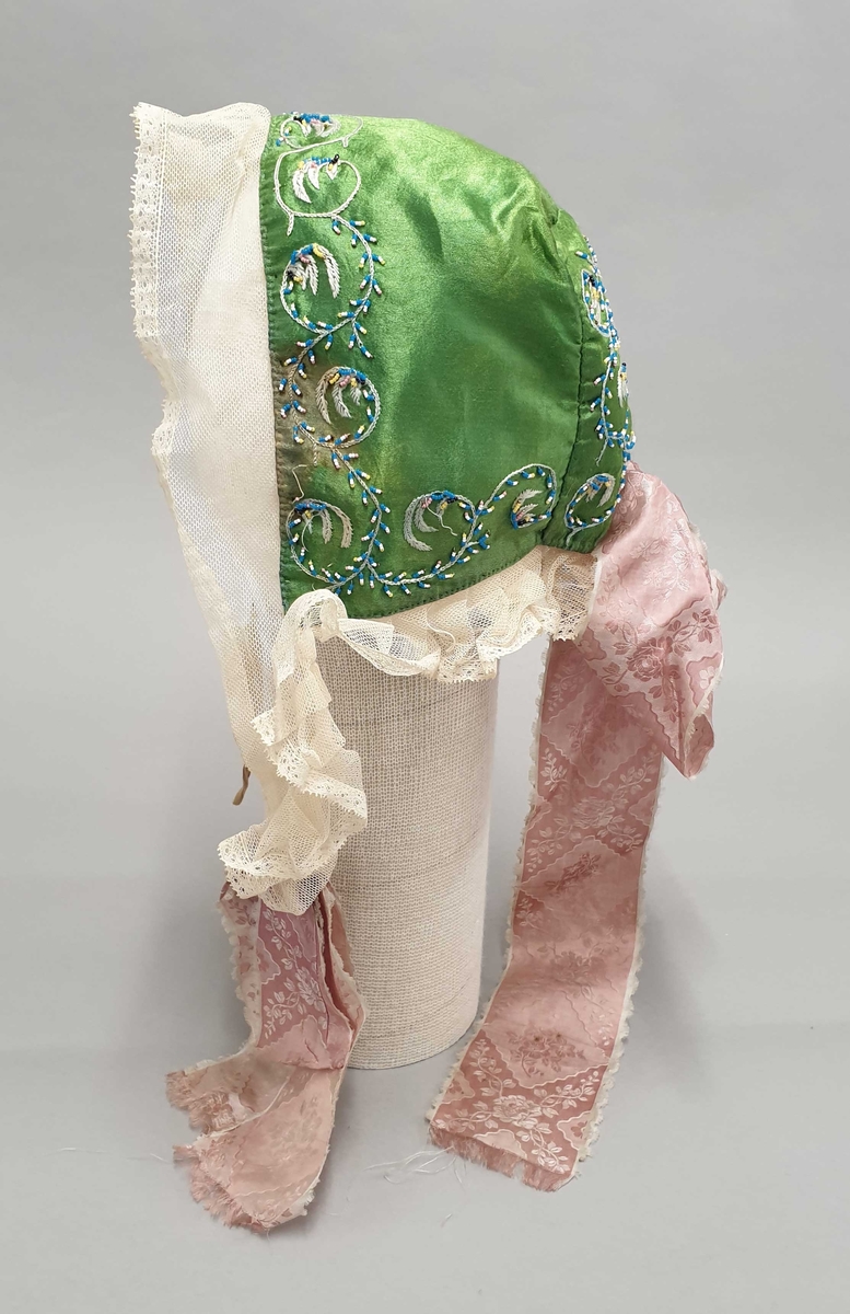 Grønn kyse av silke med brodering langs kantene og midt bak. Strimmel av hvit tyll og rosa silkesløyfe midt bak.