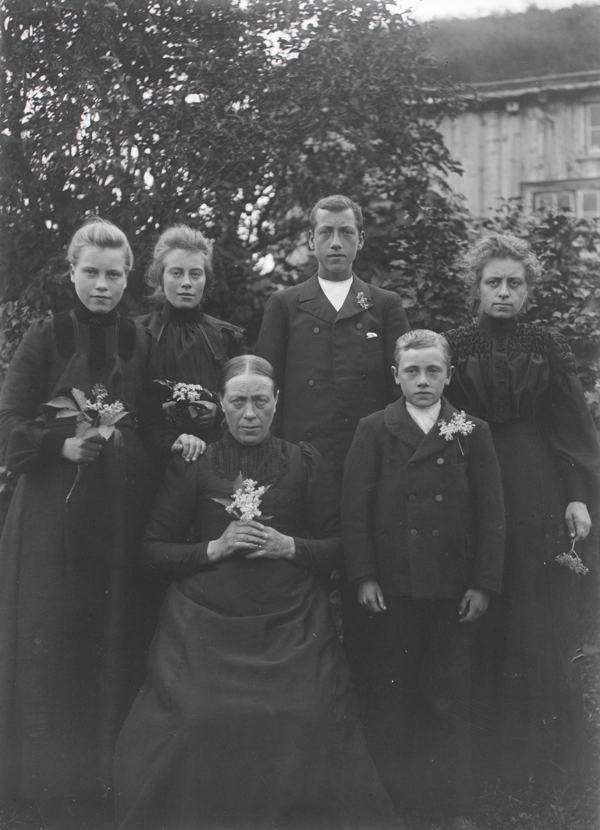 Gruppeportrett av en familie i hagen foran et hus. I hendende holder de en blomster. Familien Aston/Eikrem?