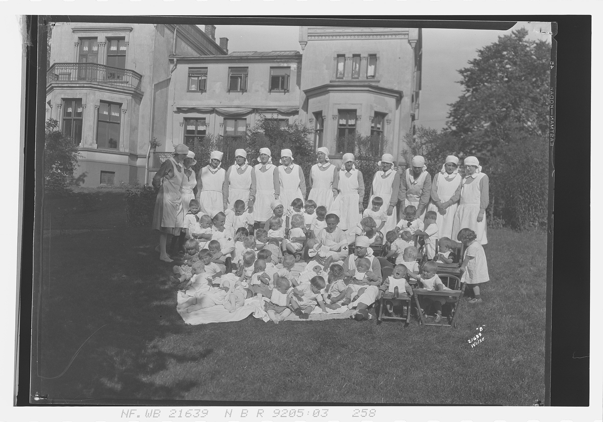 Barn og sykepleiere sitter på plenen foran Spedbarnhjemmet, Åkebergveien 23, Oslo. Fotografert 1928.