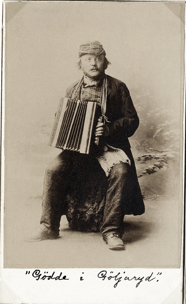Foto av en man i slitna allmogekläder med rock och mössa. Han sitter på en stubbe och spelar dragspel. 
Knäbild, halvprofil. Ateljéfoto.