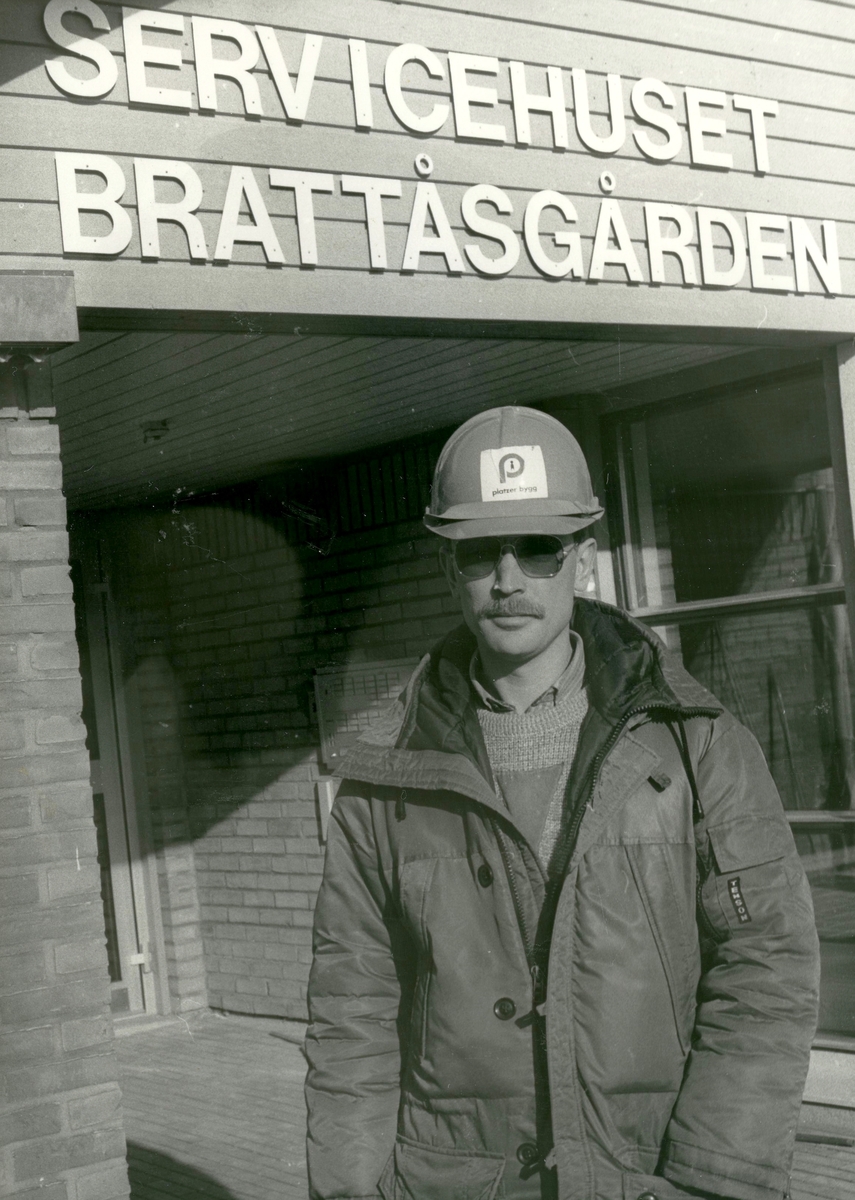 En okänd man, iklädd Tenson-jacka och med "platzer bygg-hjälm" på huvudet, står framför servicehuset Brattåsgården på Streteredsvägen 5 år 1986.