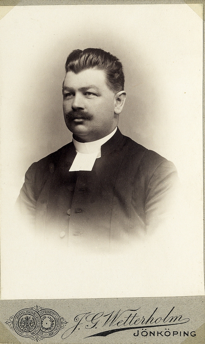 Foto av en man med mustascher, klädd i prästrock med prästkrage.
Bröstbild, halvprofil. Ateljéfoto.