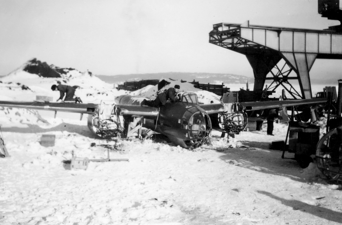 Flygtekniker jobbar med demontering av bärgat flygplan B 18 på kajen i Härnösands hamn efter nödlandning på isen den 10 februari 1946. I bakgrunden syns en lyftkran som användes vid bärgningen.