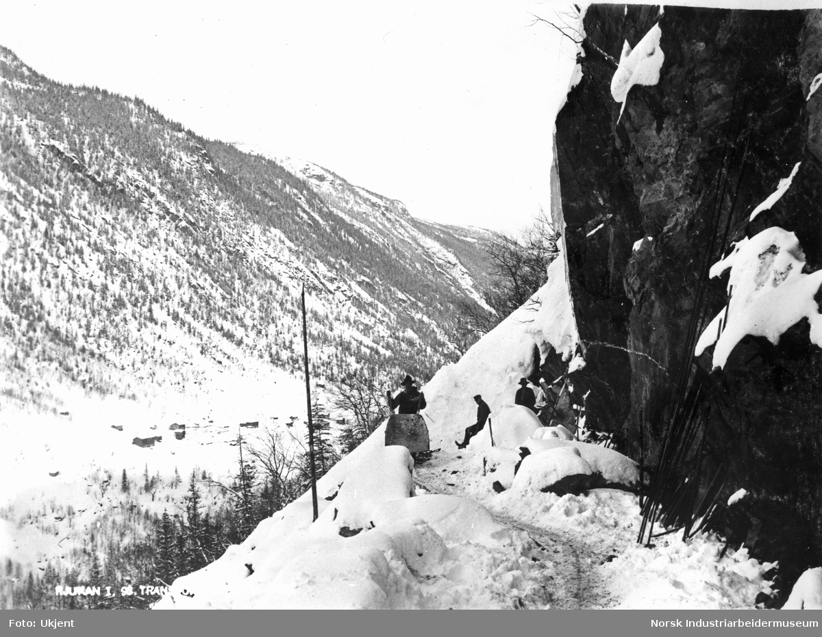 Menn i arbeid med Vemorksporet i snøen. Mann stående med trillebår. Boliger sees på andre siden av juvet langs Vesetsletta.