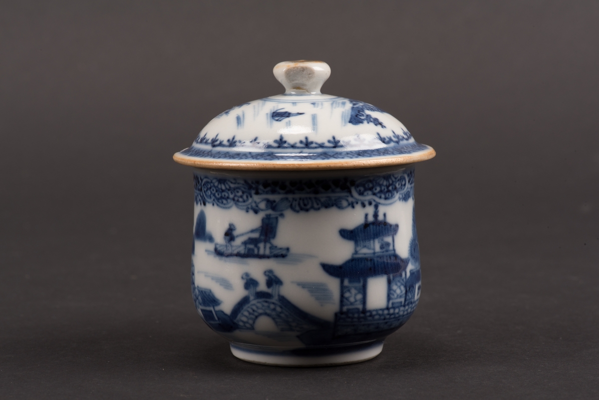 Krämkopp med lock och öra. Dekorerad med blå målning på vit botten under glasyr, med landskap och pagoder. Lockknoppen har spår efter förgyllning. Tillverkad i Kina under Qianlong-perioden.