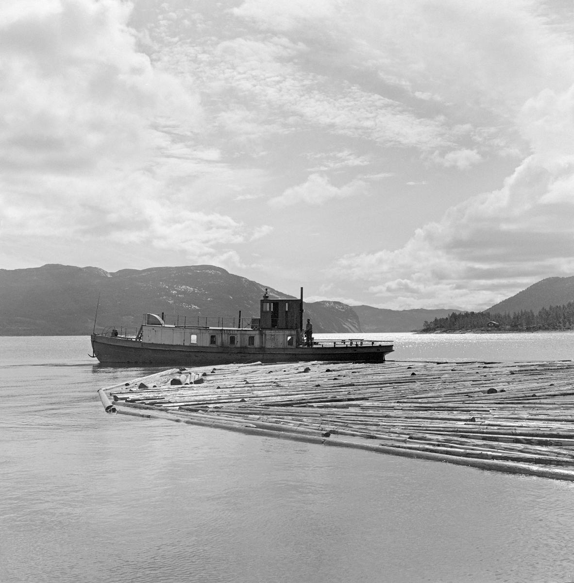 Slepebåt sleper tømmer over innsjø. Tømmersleping med slepebåten M/S Teisner på Fyresvatnet, Fyresdal, Telemark. Skipper Gunnar Sandvik . Båten ble bygd på Akers mekaniske verksted, og var opprinnelig en kombinert passasjer- og slepebåt fra 1876, Den fikk navnet M/S Teisner i 1913. Etterhvert ble det en ren slepebåt. Etter at tømmertransporten forsvant i 1971, ble båten tatt vare på av "Reiarlaget Teisners vener", men i 1990 ble båten dratt på land. Fyresvatn.