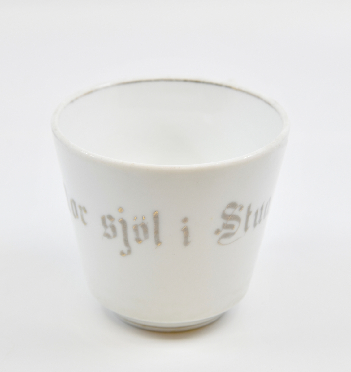 En kopp med tefat i hvitglasert porselen. Koppen har gullstripe nederst og på innsiden øverst. På koppen er det tekst i grått/gullfarge. I den ene siden er det en firkantet hank formet som en stav (stavhank).