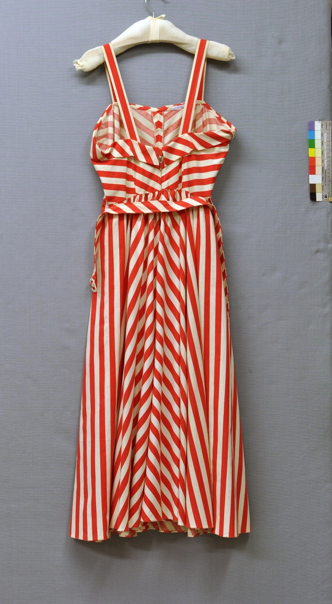 Solklänning (BM63830:1) med kortärmad jacka (BM63830:2) och skärp i röd-vitrandigt tryckt bomullstyg. Klänningen avskuren i midjan. Litet snedskuret liv med 25 mm breda axelband. Framstycket med mittsöm och 2 midjeinsnitt. Bakstycket lågt skuret med 2 midjeinsnitt. Nedvikt sneddad kant runt om. Klockad kjol av 4 våder, rynkad. Tygskärp med vitt galalitspänne. Blixtlås i ryggen.
Jackan avskuren i midjan med snedskuret skört. Framsidan med 2 bystinsnitt och 2 midjeinsnitt, knäppt med 4 vita rockknappar av galalit.  Baksidan helskuren med 2 midjeinsnitt. Skörtet med 4 lagda veck på vardera fram- och baksidan. Skjortbluskrage. Kort ärm med uppvikt snedskuret slag. Axelvaddar. Storlek 42.