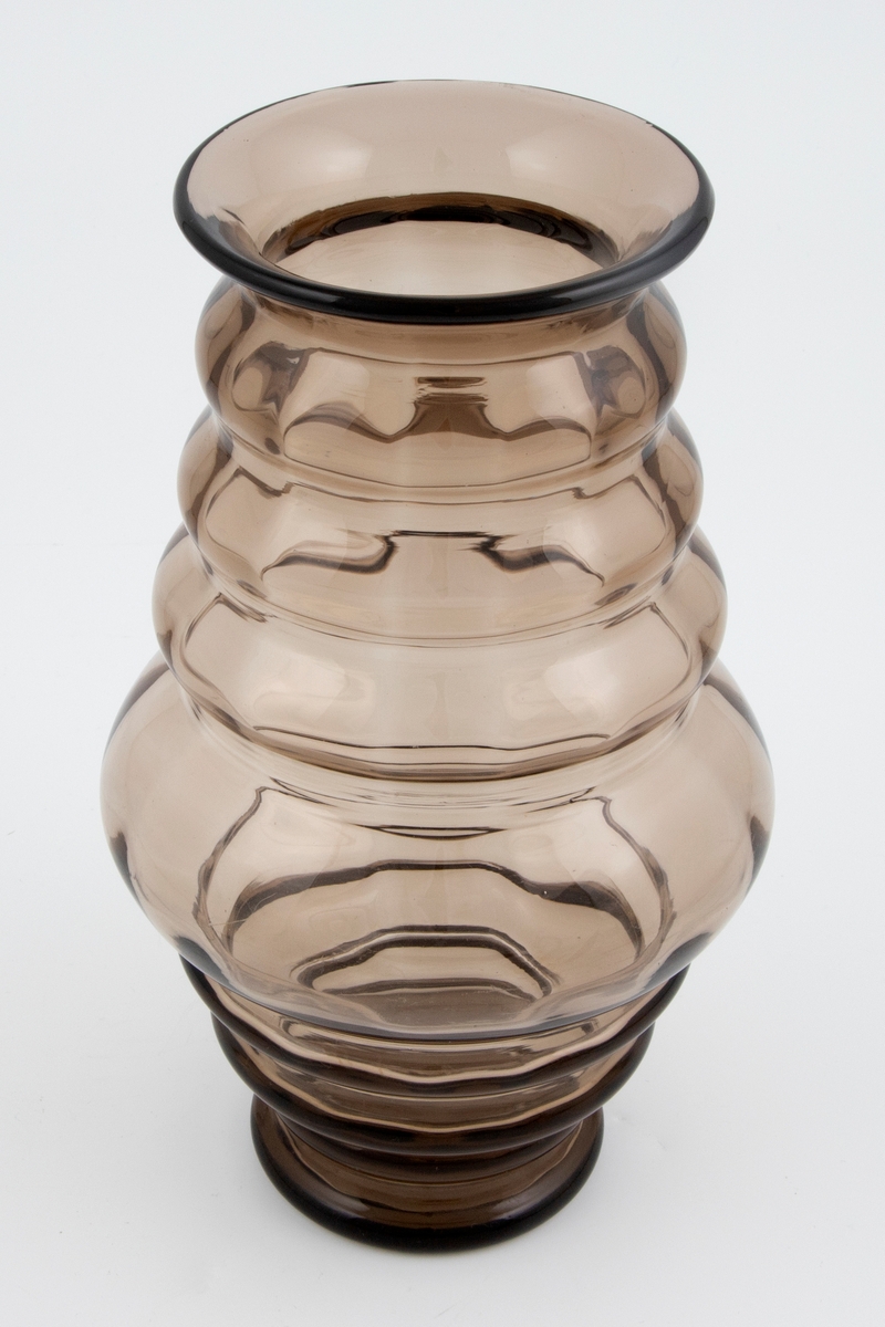 Balusterformet vase i bruntonet gjennomskinnelig glass med optisk dekor. Korpus er bygget opp av vulster som er bredest på midten av vasen, og smalner gradvis mot fot og munningsrand. Puntemerke under bunnen.